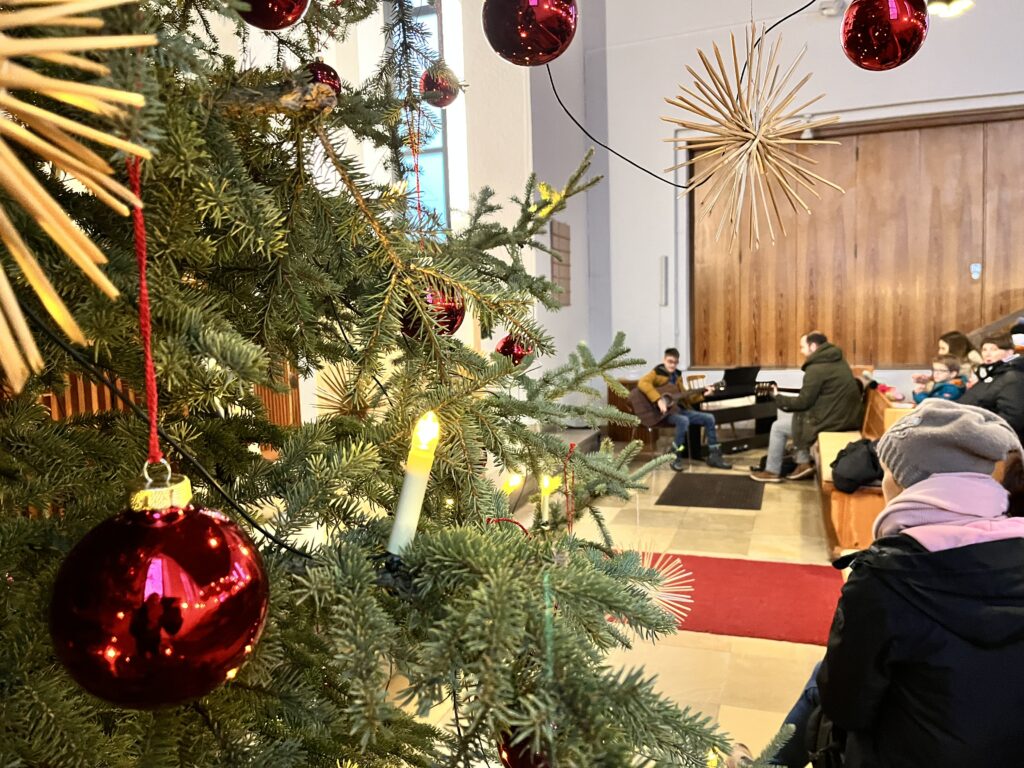 Weihnachstschmück am Weihnachtsbaum. Im Hintergrund Lehrkraft und Schüler mit Gitarre