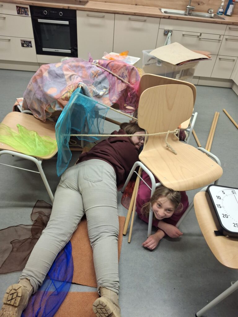 Zwei SchülerInnen liegen in einer selbstgebauten Höhle aus Stühlen und Tüschern auf dem Boden