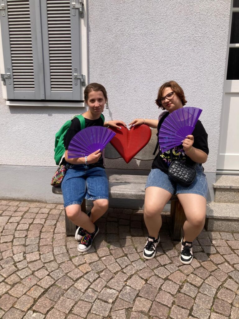 Zwei Schülerinnen sitzzen auf einer Bank mit einem Herz als Lehne zwischen ihnen